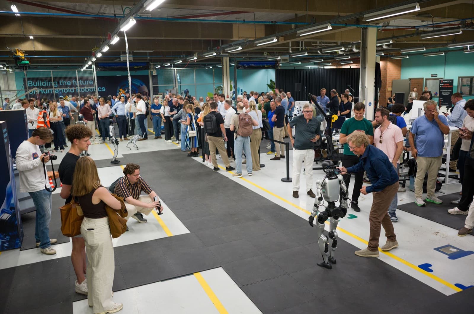 Volle opening van het HAC: bezoekers rond de humanoids op de werkvloer, onder de banner Building the future of Humanoid Robotics